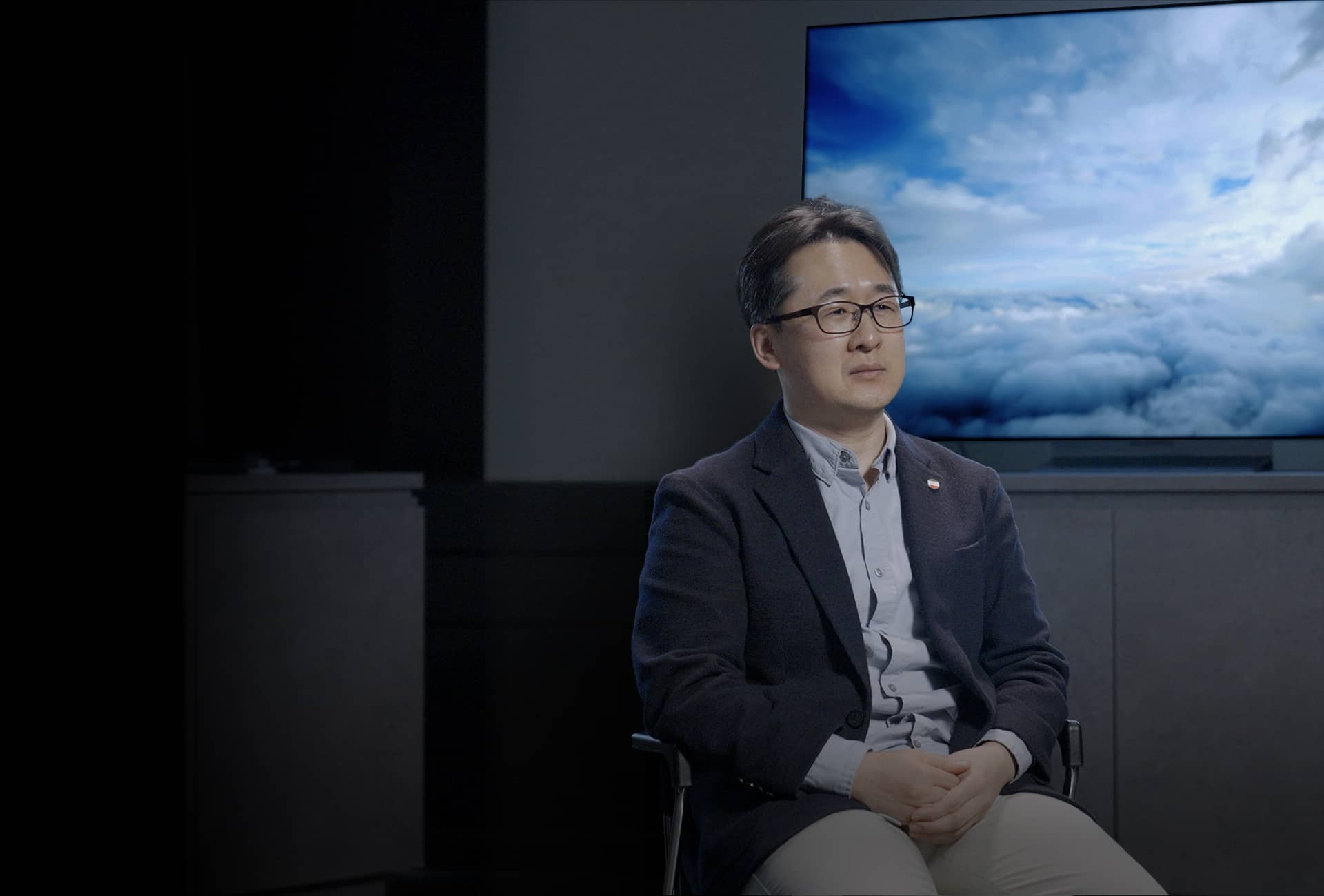 Mr.Kim is sitting in the background of the 3rd Generation META Technology OLED display, which shows cloudy skies.