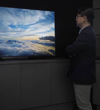 Mr.Kim is looking at META Technology OLED display with his arms crossed, and a cloudy sky image is visible on the screen.