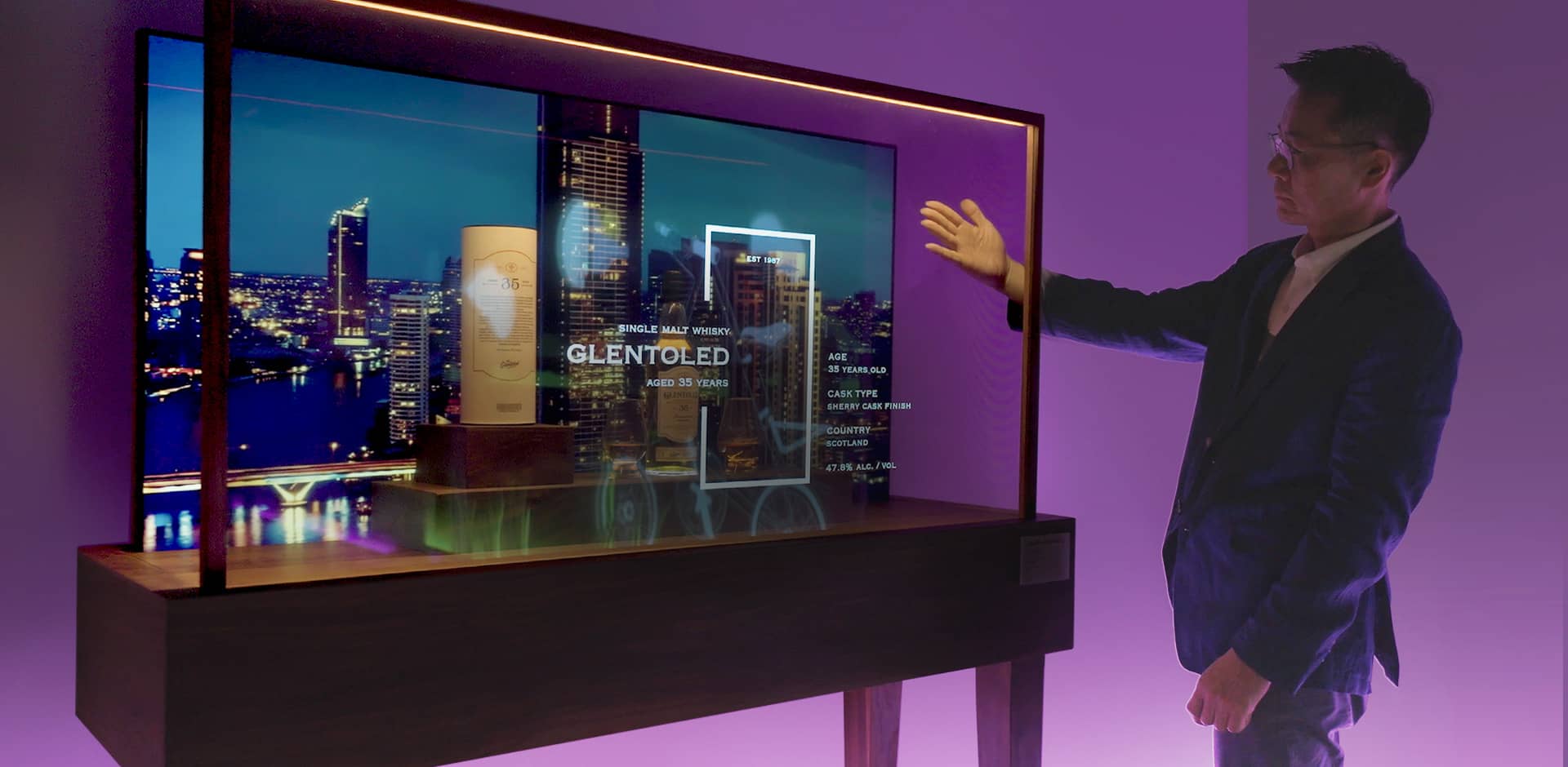 Whiskey products are displayed in the Transparent OLED showcase, and Mr. Kim is looking at the transparent screen with his hand between the displays.