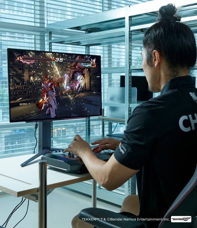 DRX 샤넬이 게이밍 OLED 모니터를 보며 철권 게임을 하고 있고, 화면에는 격투 장면이 보인다.