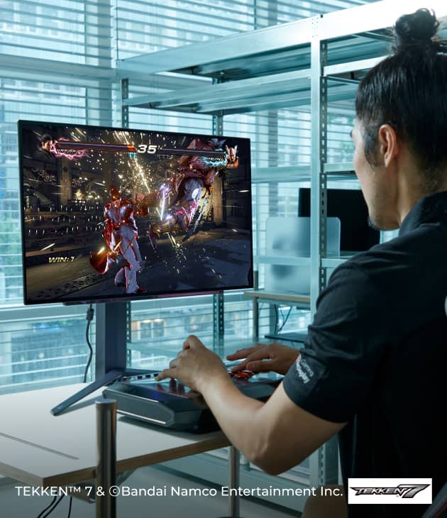 DRX 샤넬이 게이밍 OLED 모니터를 보며 철권 게임을 하고 있고, 화면에는 격투 장면이 보인다.